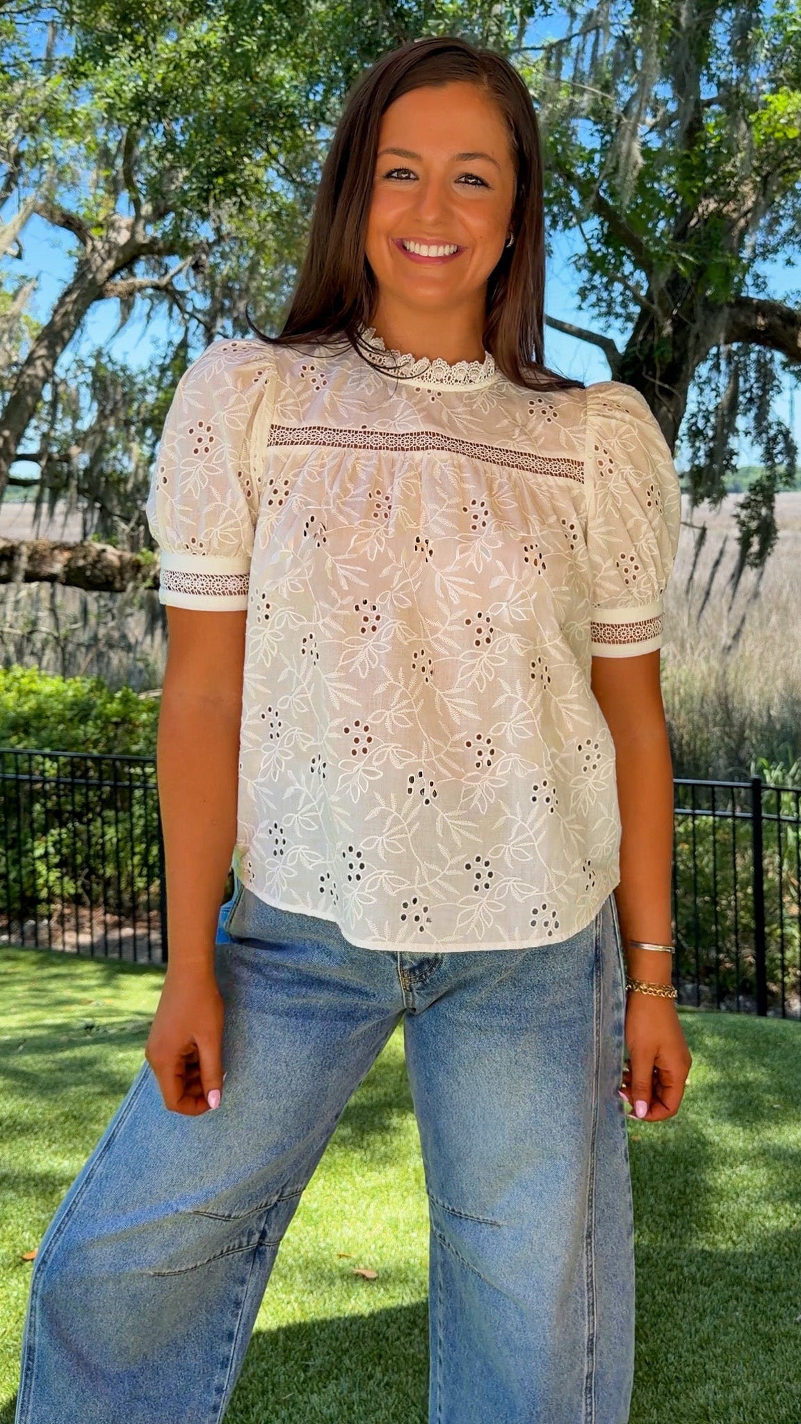 Woman wearing a white lace top and blue jeans standing outdoors with trees and grass in the background