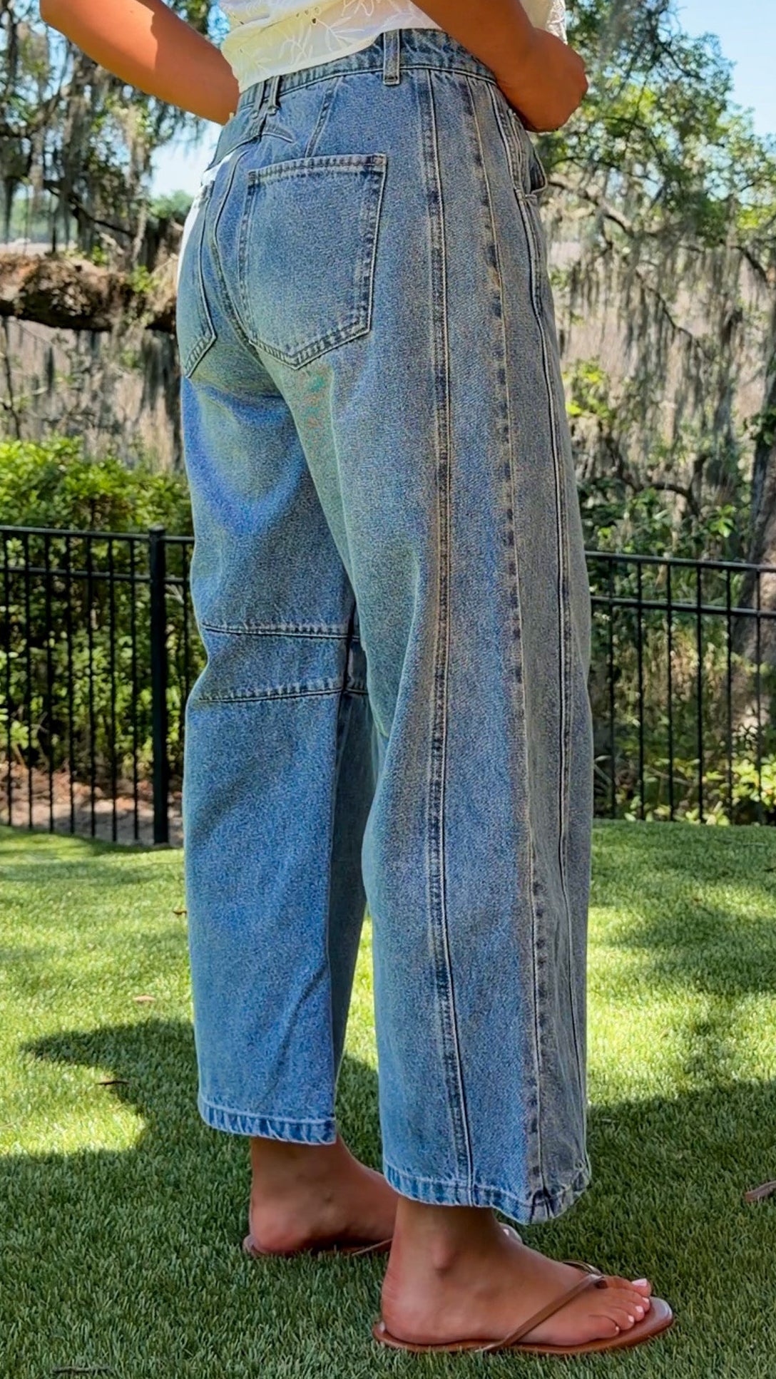 Person wearing light blue jeans standing on grass with trees and a fence in the background