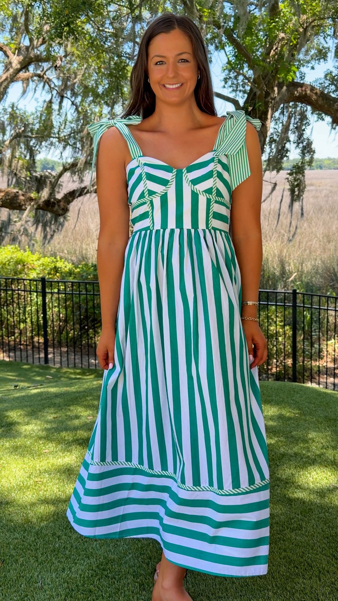 Woman wearing a green and white striped dress standing outdoors with trees and grass in the background