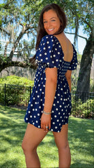 Woman in a blue polka dot dress standing outdoors with trees and grass in the background