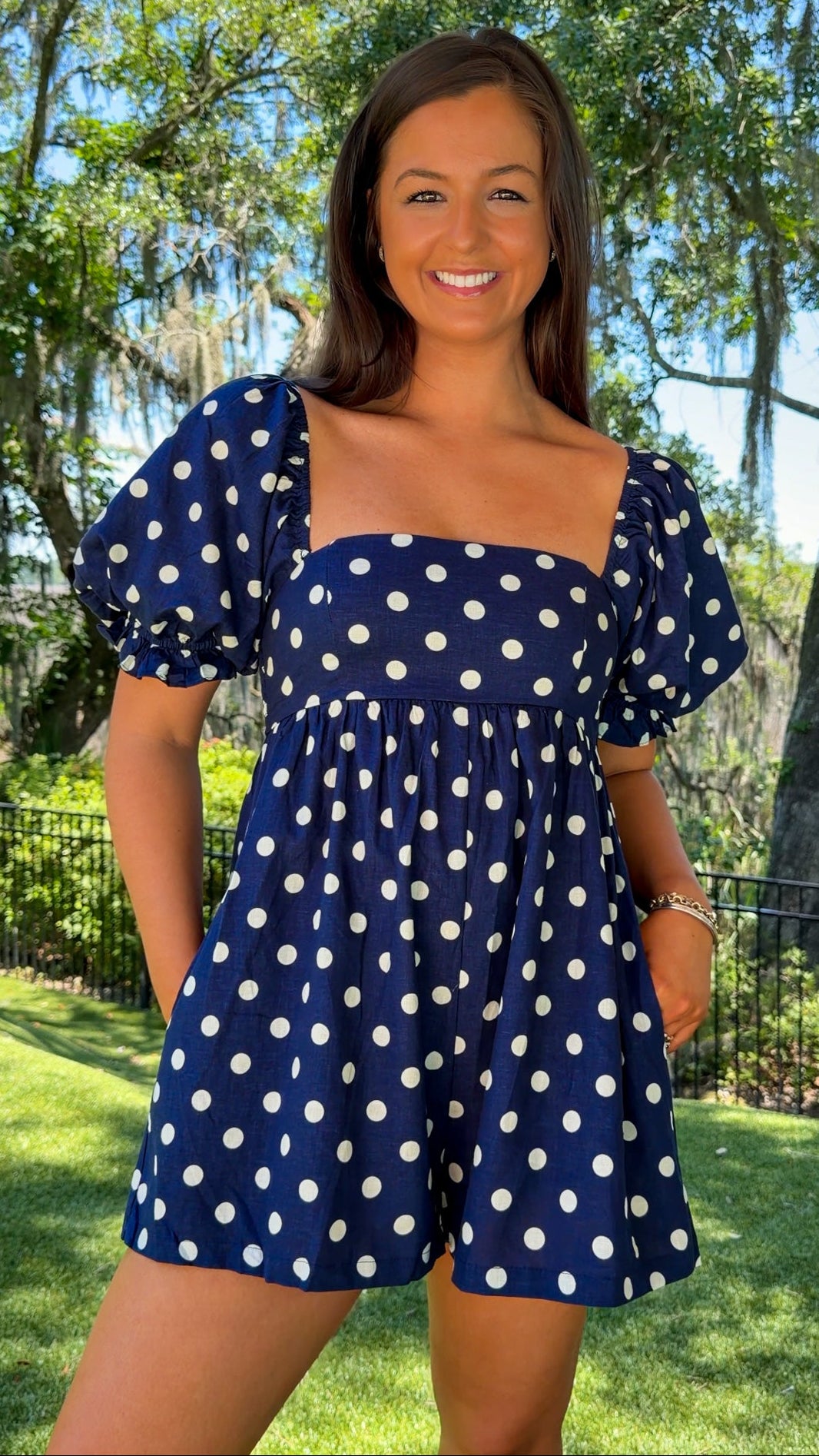 Woman wearing a blue polka dot dress standing outdoors with trees in the background