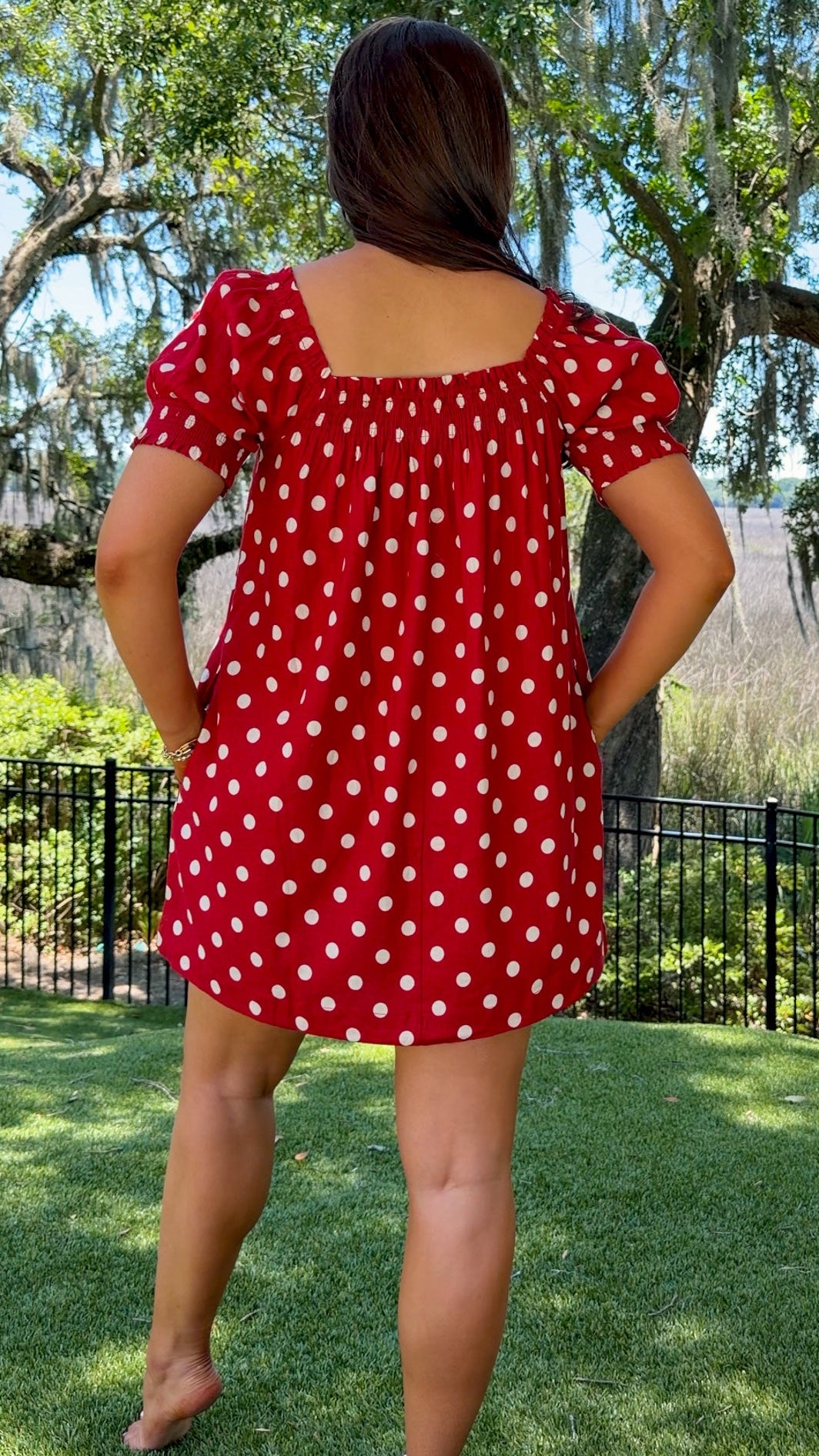 Person wearing a red polka dot dress standing outdoors with trees and grass in the background