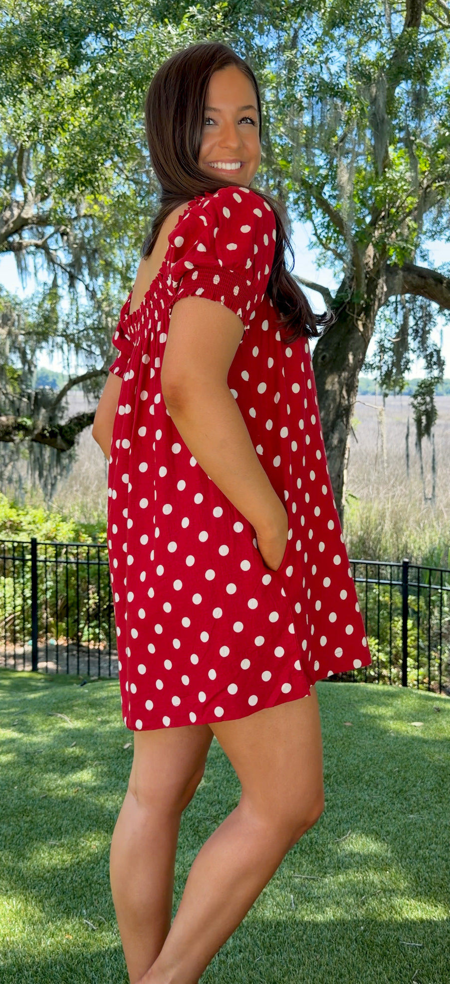 Woman wearing a red polka dot dress standing outdoors with trees and grass in the background