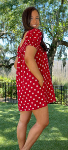 Woman wearing a red polka dot dress standing outdoors with trees and grass in the background