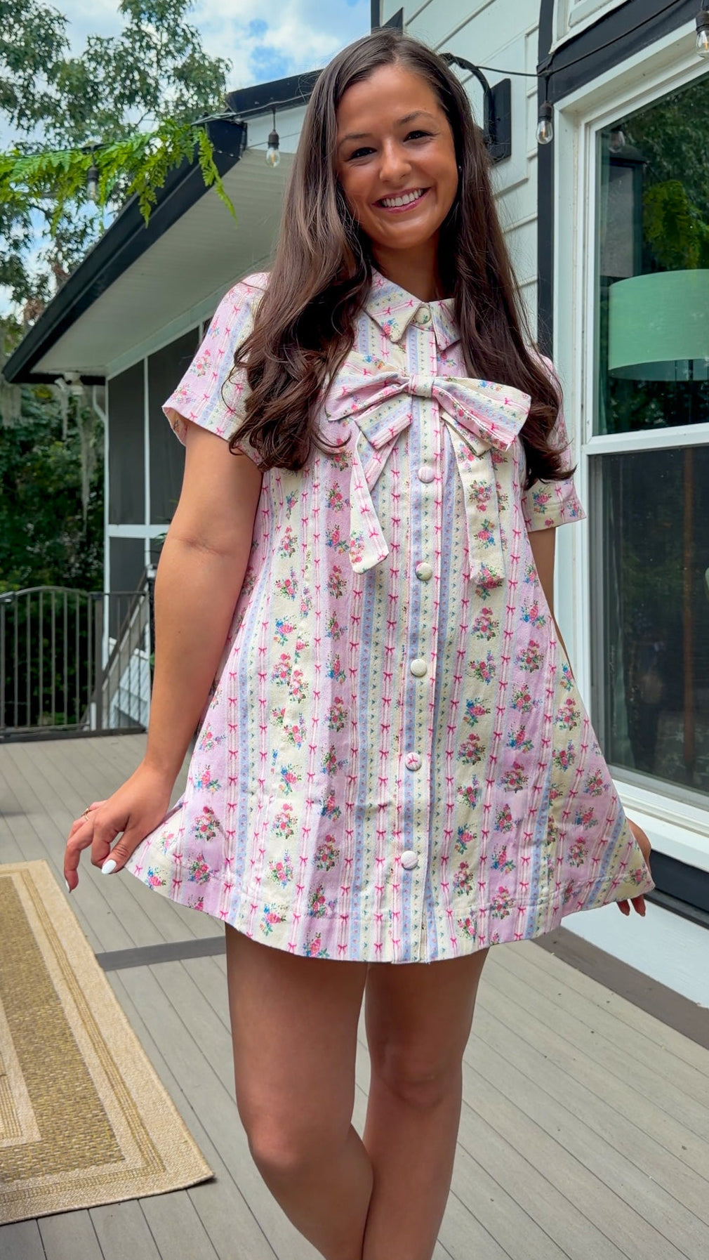 Light pink floral stripe mini dress with a collared neckline, front button closure, short sleeves, oversized bow detail, and relaxed A-line silhouette.