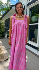 A woman wearing a pink and white vertically striped sleeveless maxi dress with tie-shoulder straps, standing on a wooden deck in front of a house.