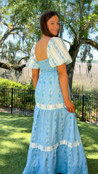 Woman in a blue floral dress standing outdoors with trees and grass in the background