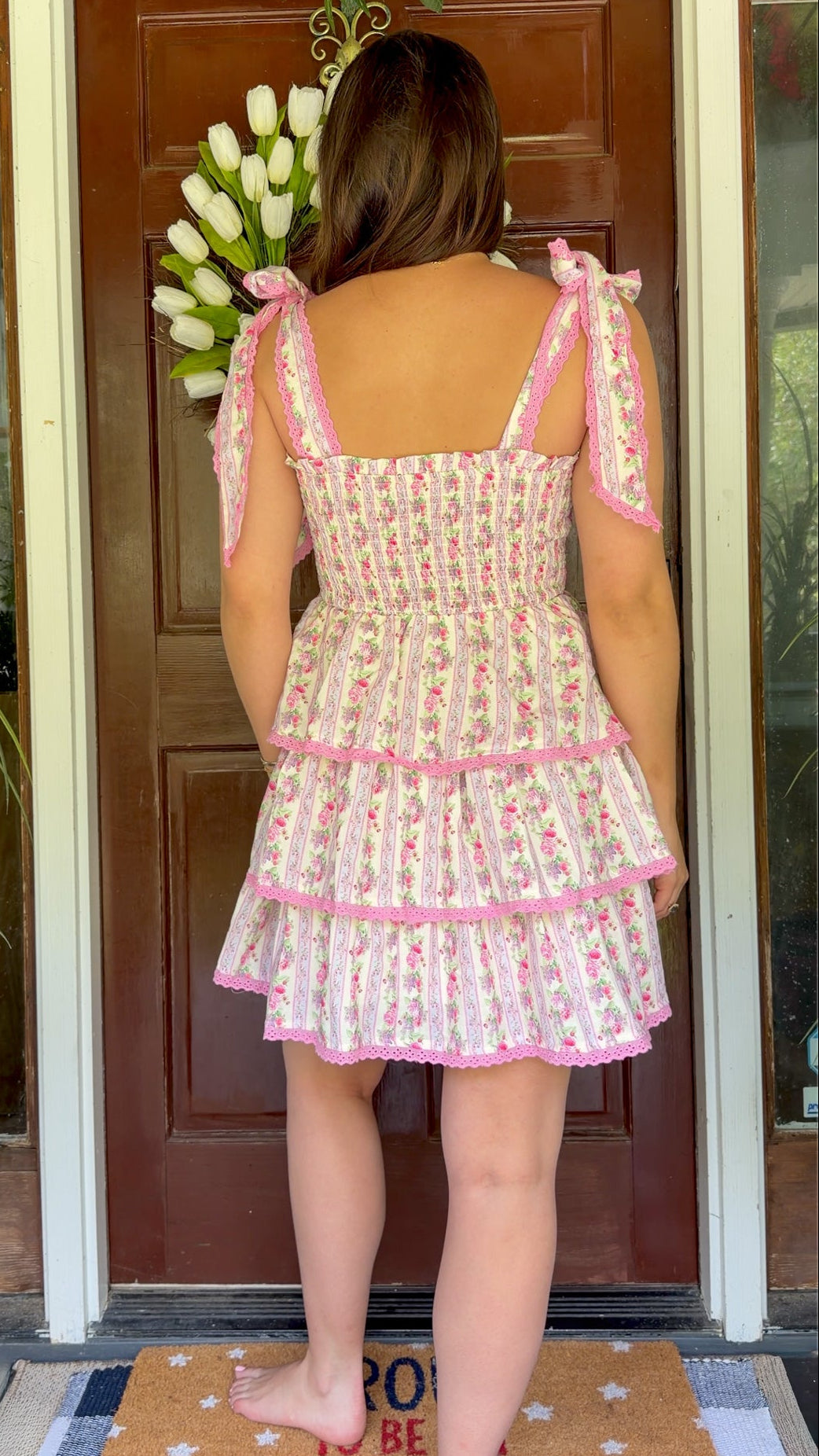 Pastel pink floral tiered mini dress with a smocked bodice, adjustable shoulder tie straps, lace trim detailing, and a flowy tiered skirt.