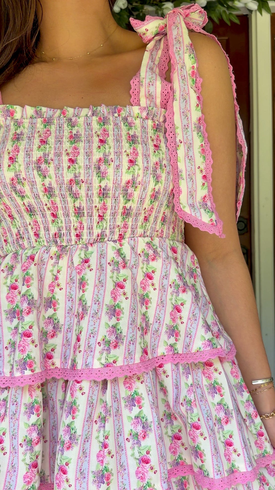 Pastel pink floral tiered mini dress with a smocked bodice, adjustable shoulder tie straps, lace trim detailing, and a flowy tiered skirt.