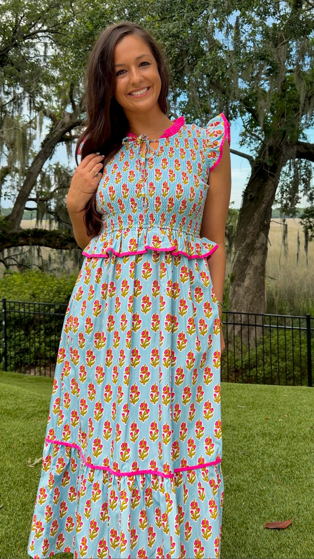 Light blue floral print midi dress featuring a fully smocked bodice, flutter sleeves with pink contrast trim, a keyhole tie neckline, ruffled peplum waist, tiered midi skirt, and side pockets.