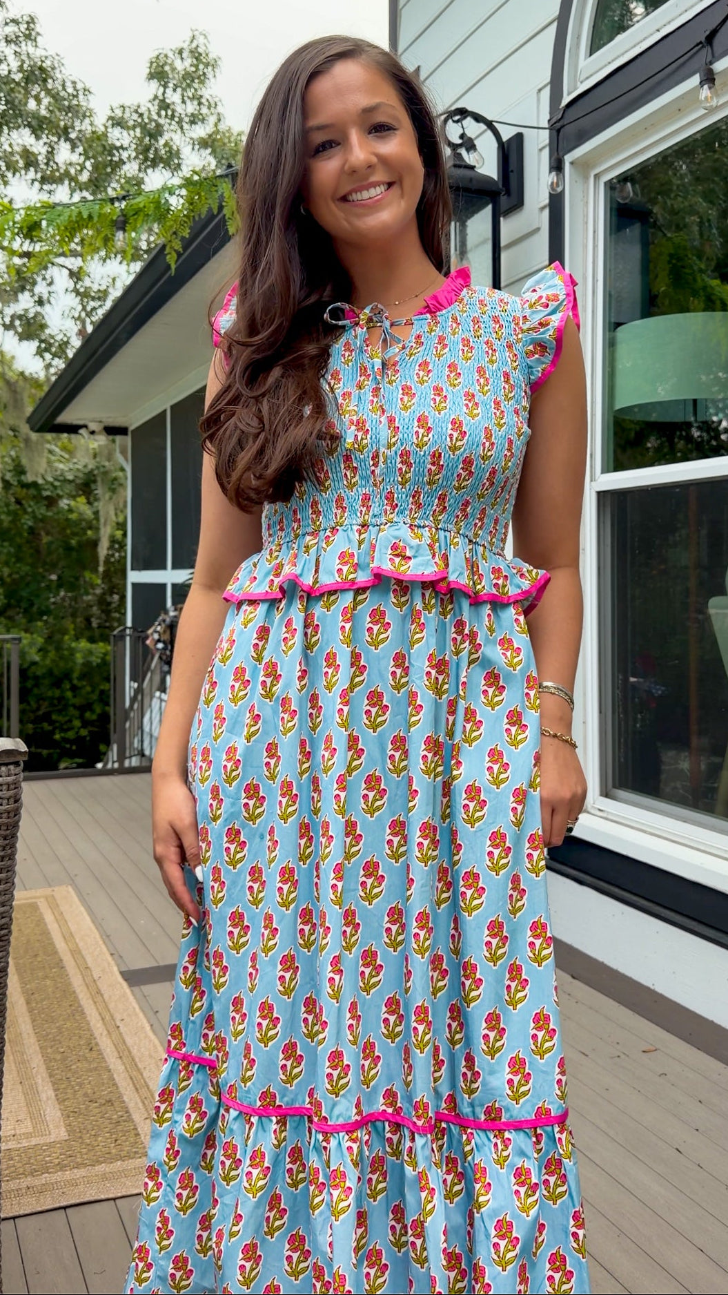 A woman wearing a light blue maxi dress with a colorful floral print and bright pink ruffle trim, standing on a wooden deck in front of a house.