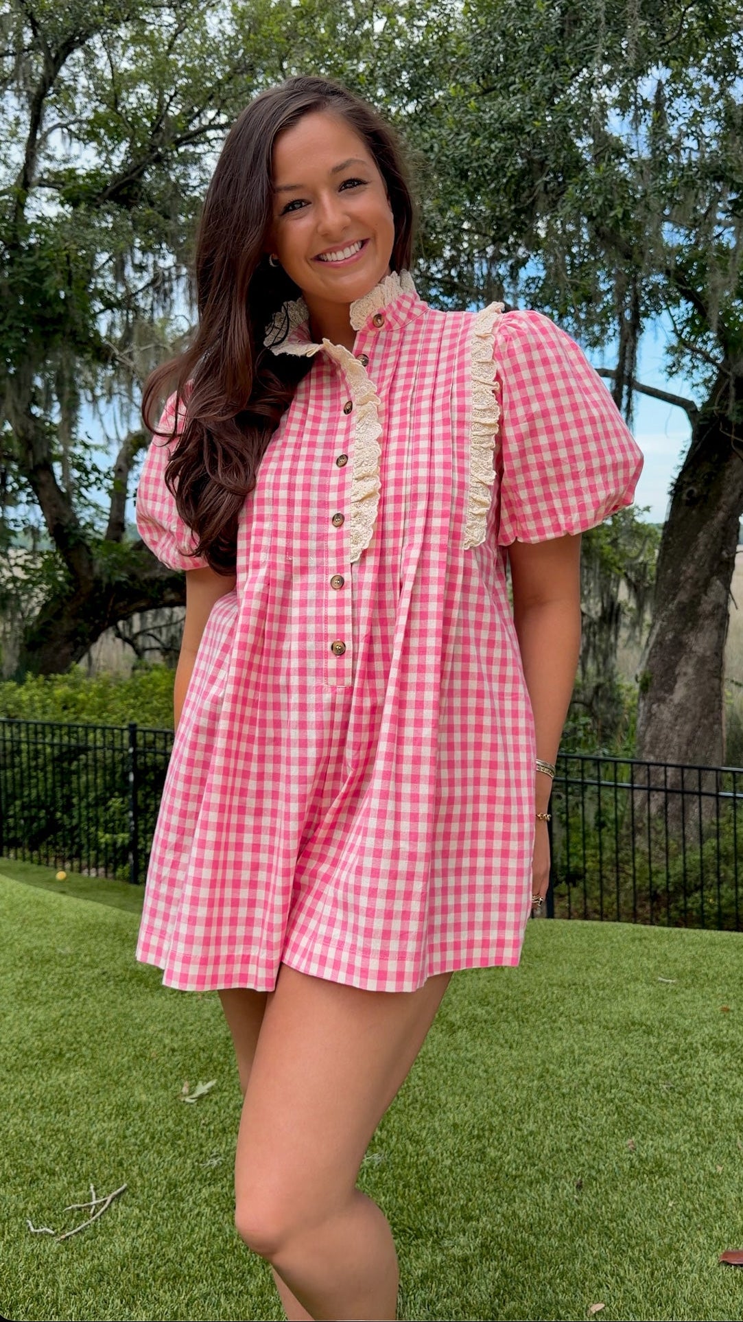 A woman wearing a pink and white gingham short-sleeve mini dress with cream ruffle trim and buttons, standing outdoors on a green lawn with trees in the background.