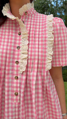 A close-up of a person wearing a pink and white gingham dress with cream lace trim and brown buttons, photographed outdoors with trees and grass in the background.