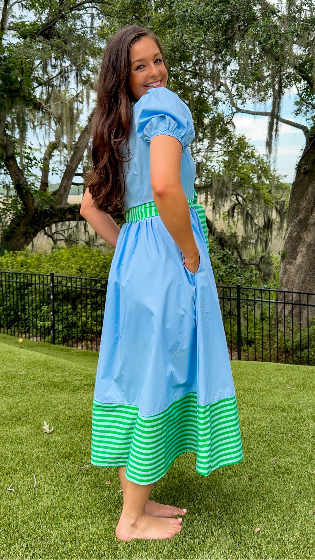 A woman wearing a light blue midi dress with green and white striped accents at the waist and hem, standing barefoot on a green lawn with trees in the background.