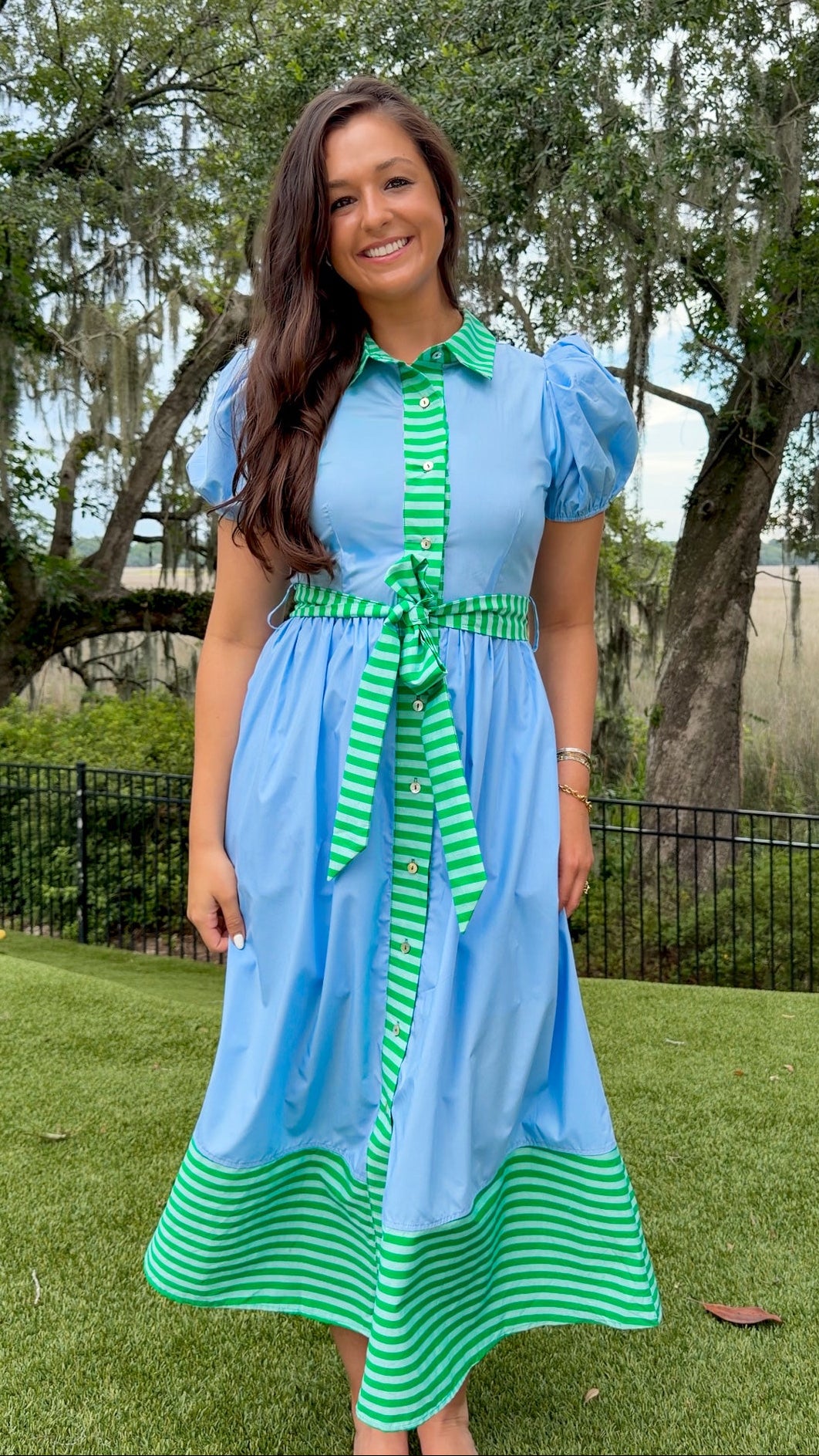 Sky blue midi dress featuring green striped contrast trim, a collared button-front design, puff sleeves, removable self-tie waist sash, side pockets, and a flowing midi-length skirt.