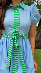Sky blue midi dress featuring green striped contrast trim, a collared button-front design, puff sleeves, removable self-tie waist sash, side pockets, and a flowing midi-length skirt.