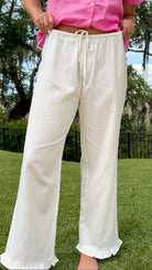 White full-length straight-leg pants featuring an elastic drawstring waistband, side and back patch pockets, delicate ruffle trim at the hem, and a relaxed, lightweight fit.
