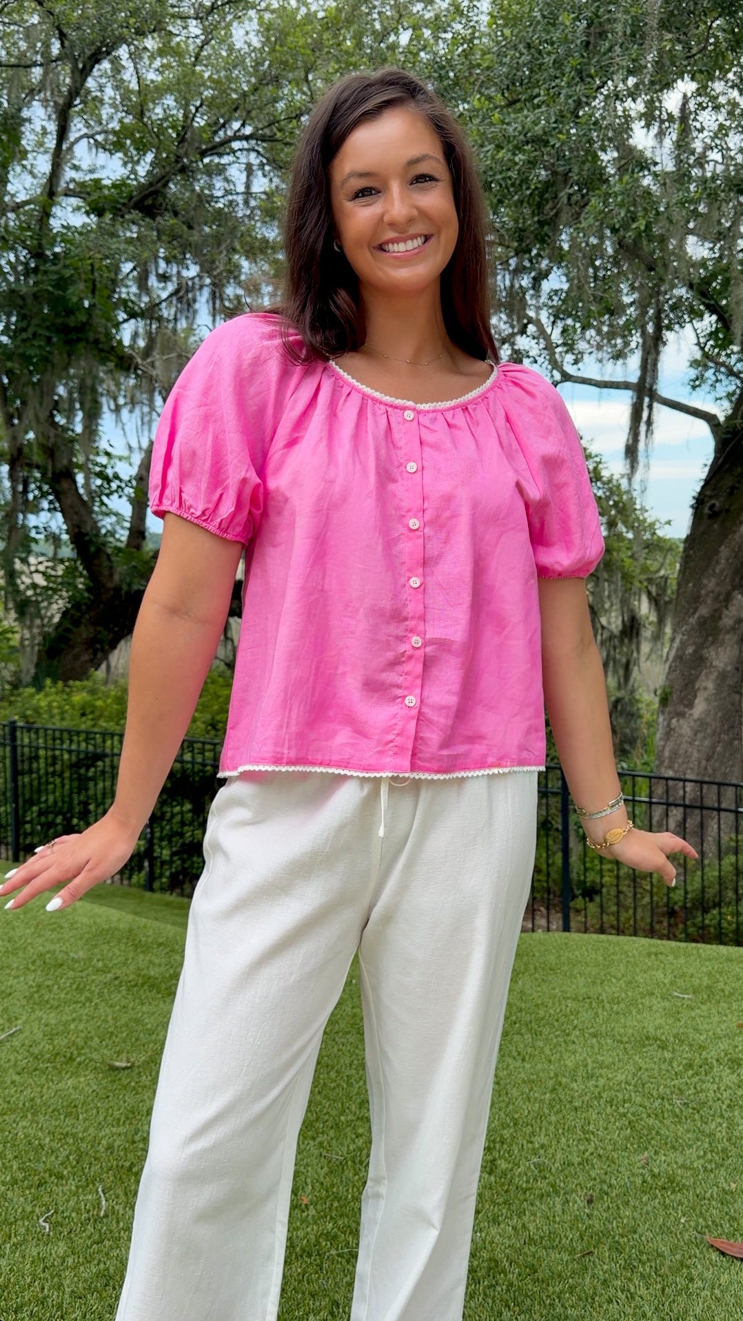 A woman wearing a bright pink short-sleeve puff-sleeve top with white buttons and white wide-leg pants, standing outdoors on a green lawn with trees in the background.