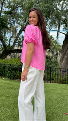 A woman wearing a bright pink puff-sleeve top and white wide-leg pants, standing on a grassy lawn with trees and a fence in the background.