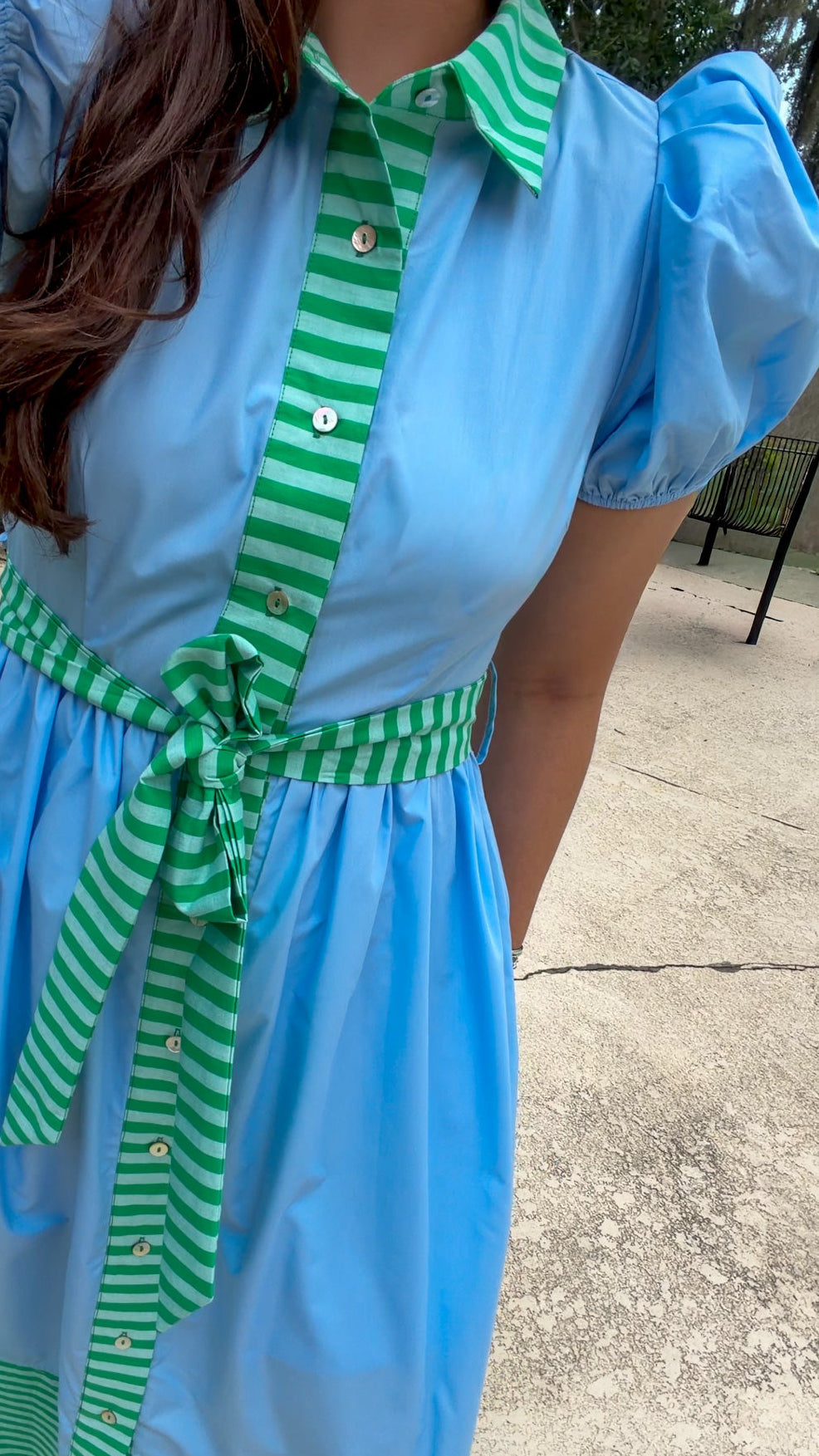 A woman wearing a light blue short-sleeve shirt dress with green and white striped trim on the collar, front placket, waist tie, and hem, standing outdoors on a concrete surface.