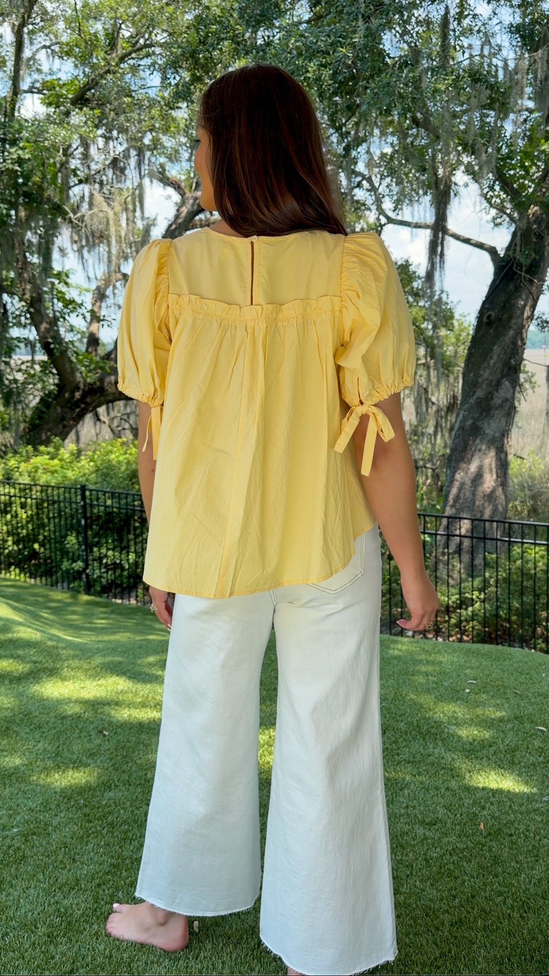 Butter yellow babydoll top featuring a round neckline, tiered shirring, short puff sleeves with adjustable tie cuffs, a ruffle-trimmed back, keyhole button closure, and a relaxed semi-cropped silhouette.