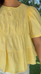 Butter yellow babydoll top featuring a round neckline, tiered shirring, short puff sleeves with adjustable tie cuffs, a ruffle-trimmed back, keyhole button closure, and a relaxed semi-cropped silhouette.