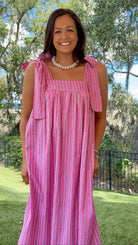 A woman wearing a pink and white vertically striped sleeveless maxi dress with tie-shoulder straps, standing outdoors in a grassy park area with trees and a fence in the background.