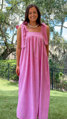 A woman wearing a pink and red vertically striped maxi dress with tie-shoulder straps, standing outdoors on a grassy lawn with trees and a fence in the background.