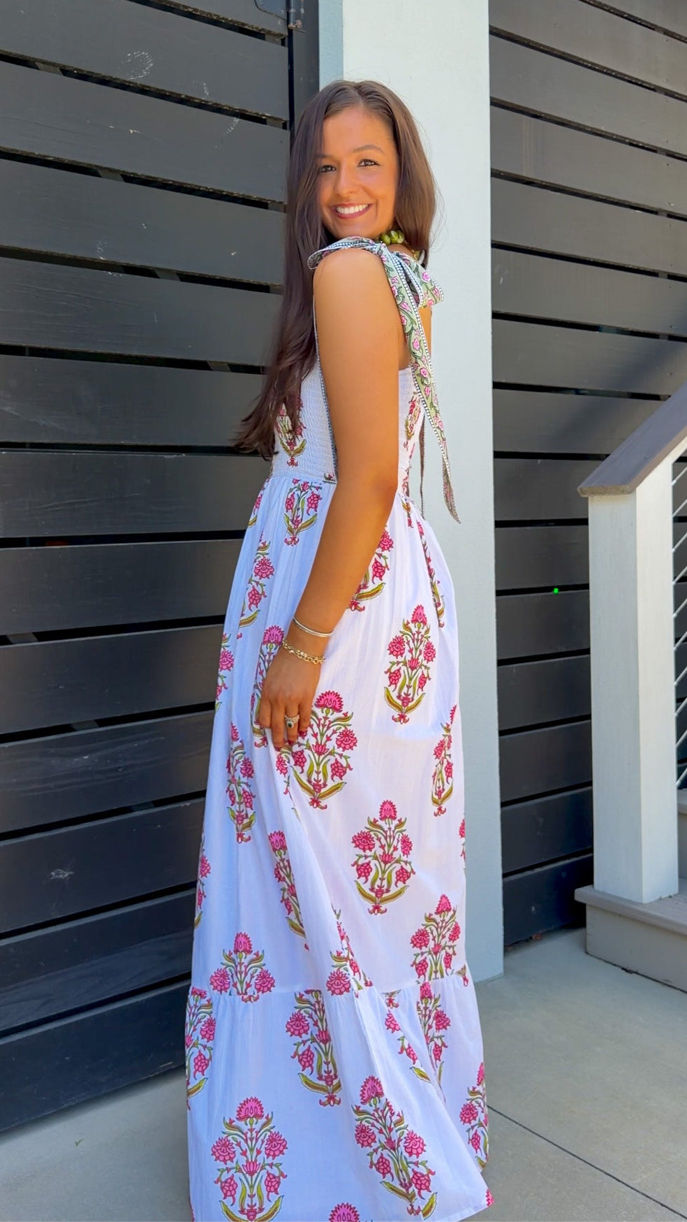 A woman wearing a sleeveless white maxi dress with pink and gold floral block prints, standing in front of a dark gray wooden slat wall.