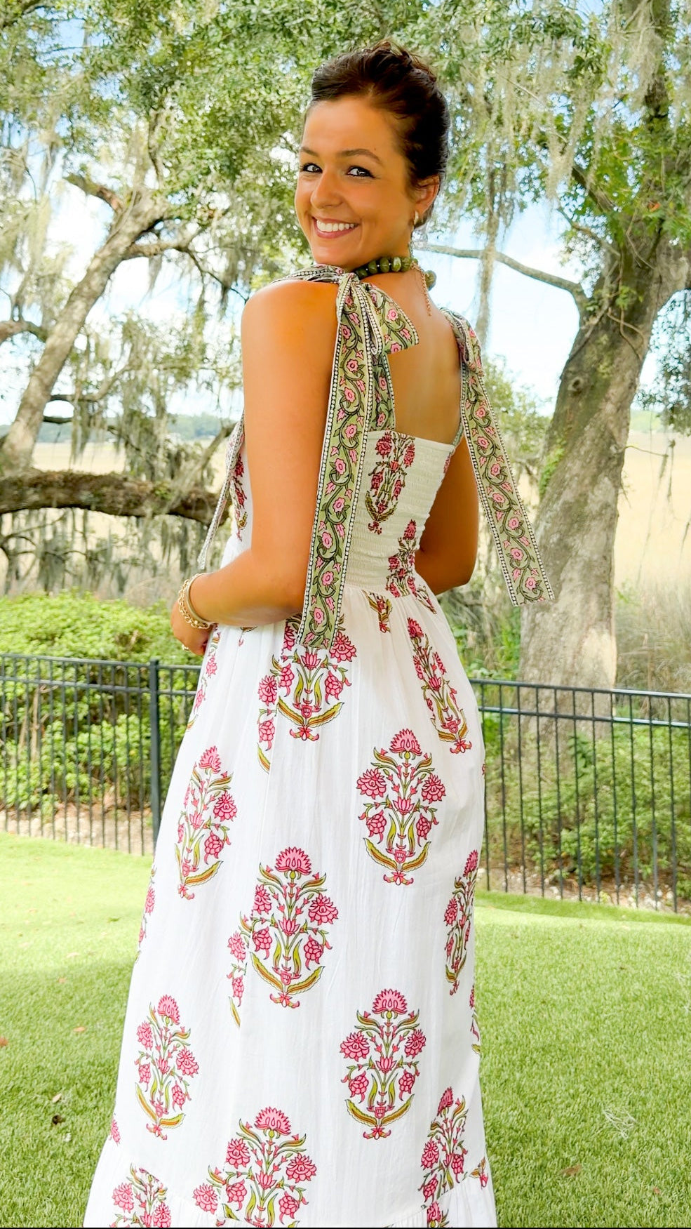 A woman wearing a white sleeveless maxi dress with pink and green floral block prints, photographed outdoors in a park with trees and grass in the background.