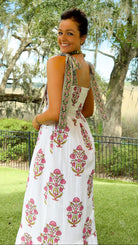 A woman wearing a white sleeveless maxi dress with pink and green floral block prints, photographed outdoors in a park with trees and grass in the background.
