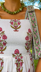 A close-up of a person wearing a white smocked dress with pink and green floral embroidery and wide decorative shoulder straps, photographed outdoors with trees and grass in the background.