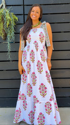A woman wearing a sleeveless white maxi dress with pink and green floral block prints stands in front of a dark gray wooden slat door.