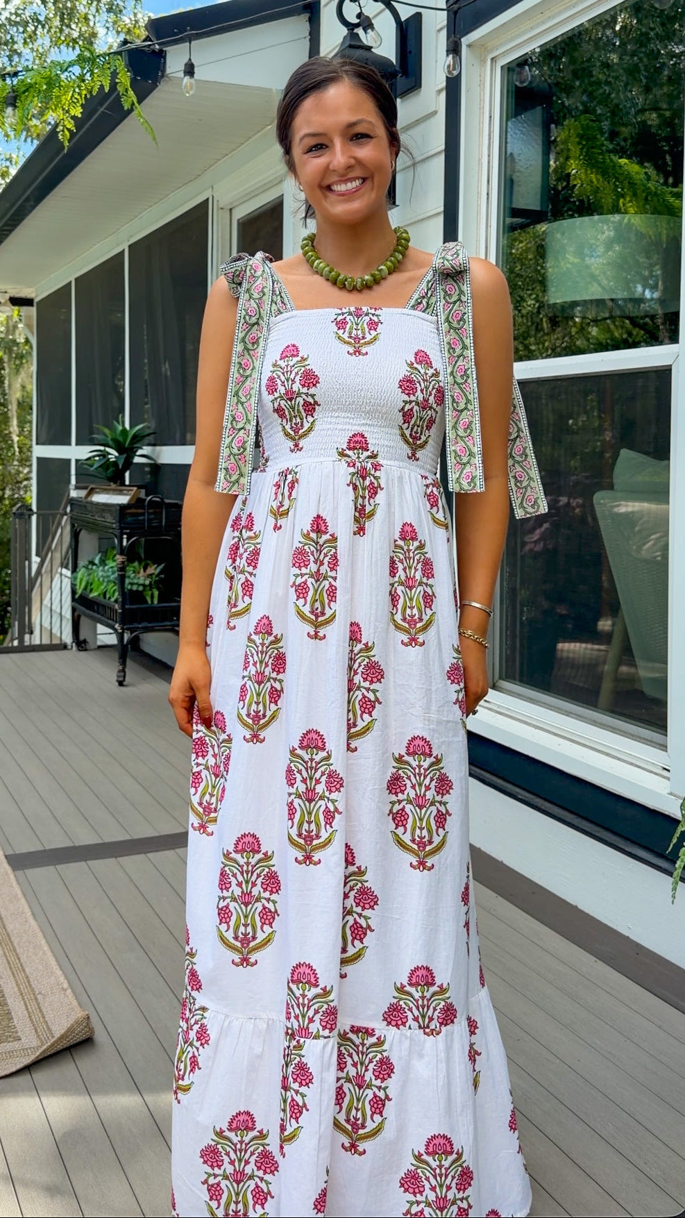A woman stands on a house deck wearing a white sleeveless maxi dress with pink and green block prints and sheer patterned flutter sleeves. The background shows a modern house exterior with large windows, screened porch area, and green trees under a blue sky.