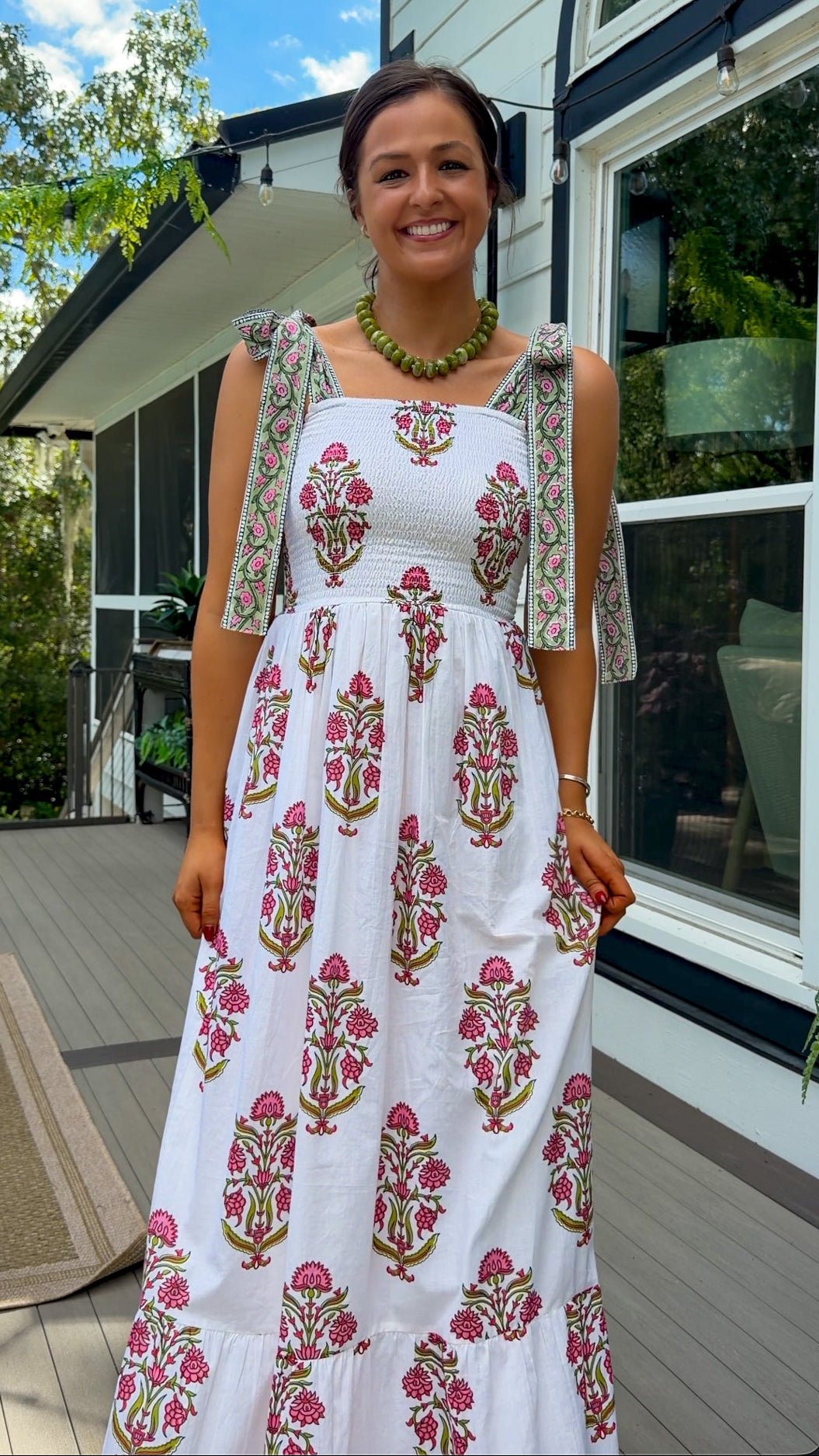 A woman stands on a wooden deck wearing a white sleeveless maxi dress with pink and green floral block prints and matching printed shoulder ties. The background shows a modern house exterior with large windows, green trees, and blue sky.