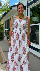 A woman stands on a wooden deck wearing a white sleeveless maxi dress with pink and green floral block prints and matching printed shoulder ties. The background shows a modern house exterior with large windows, green trees, and blue sky.