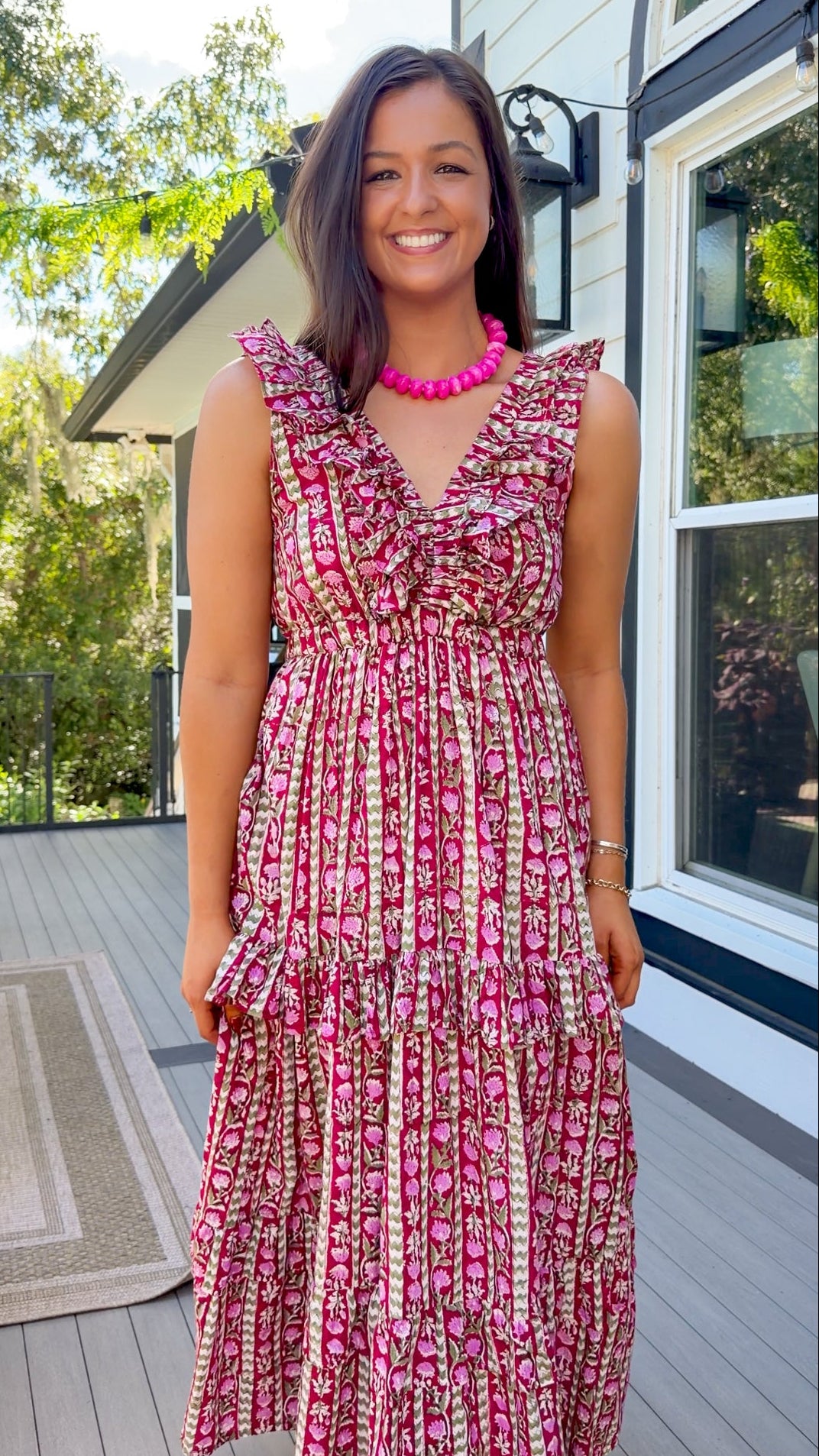 A woman wearing a sleeveless tiered maxi dress with a pink, red, and green floral pattern, standing on a wooden deck in front of a house.