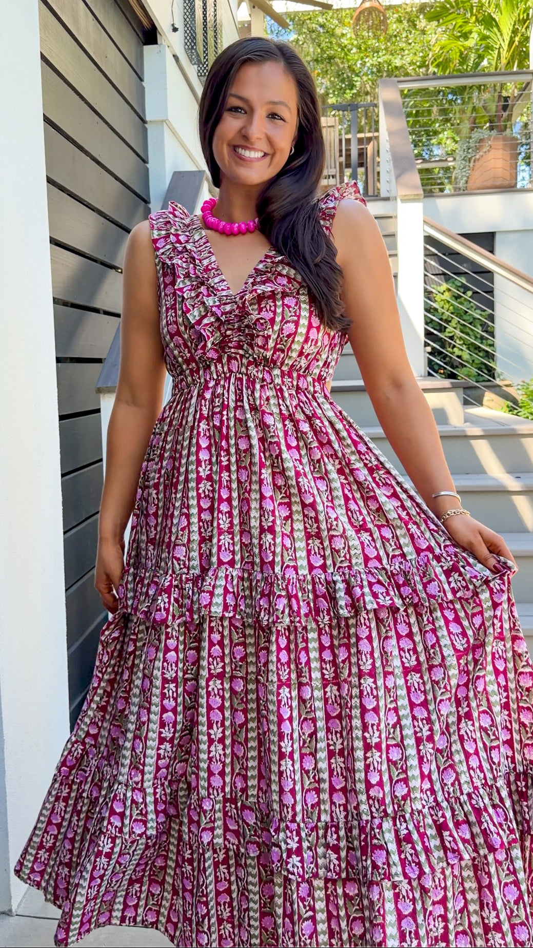 A woman wearing a tiered sleeveless maxi dress with a pink, brown, and white patterned print, standing outdoors in front of a modern house with gray siding and a staircase.