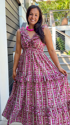 A woman wearing a tiered sleeveless maxi dress with a pink, brown, and white patterned print, standing outdoors in front of a modern house with gray siding and a staircase.