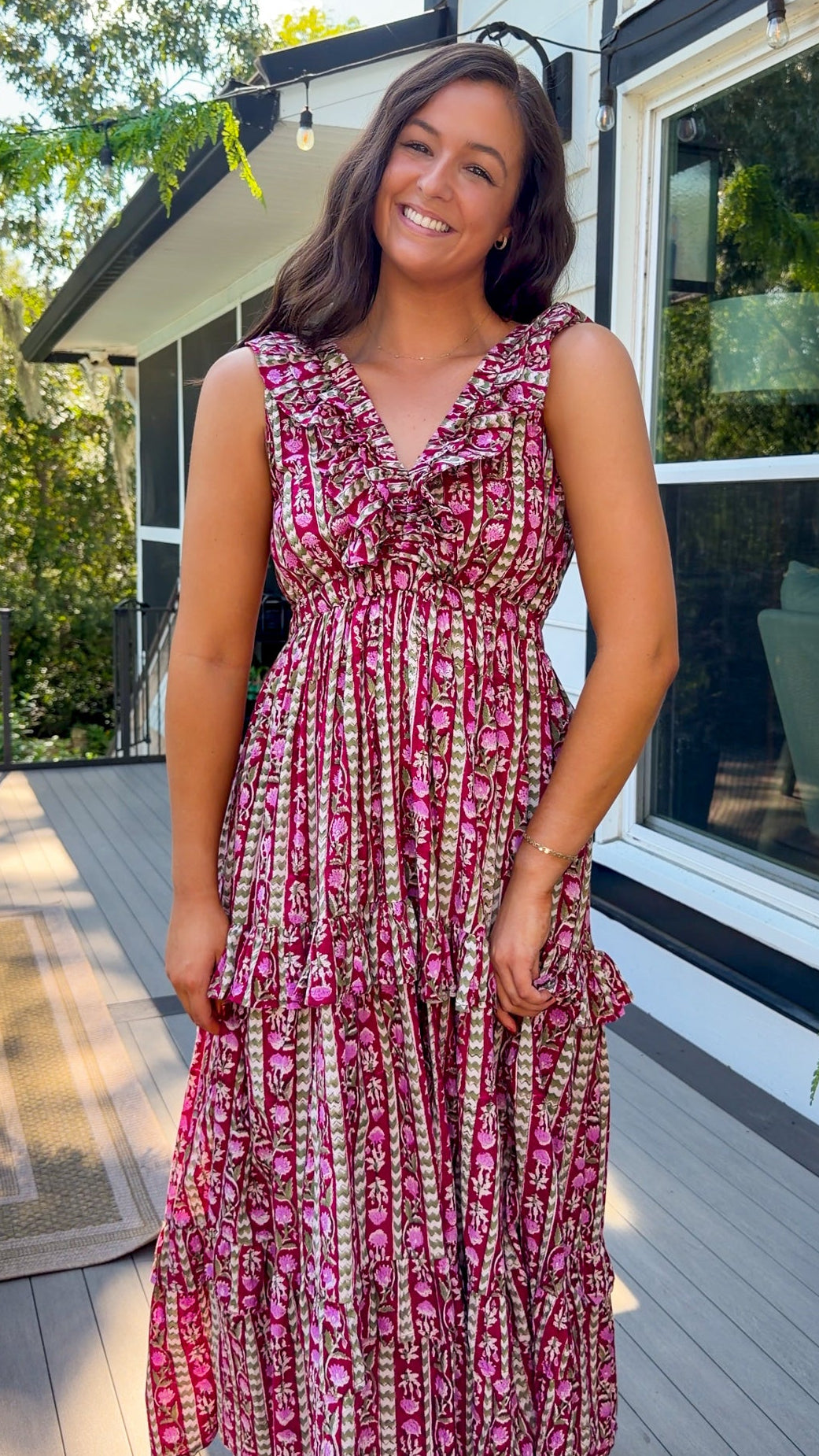 A woman wearing a sleeveless tiered maxi dress with a pink, red, and green floral pattern, standing on a wooden deck in front of a house.